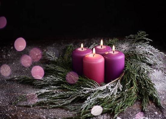 Christmas eve, wreath with four burning purple advent candles on a dark wooden snowy background with festive bokeh lights.